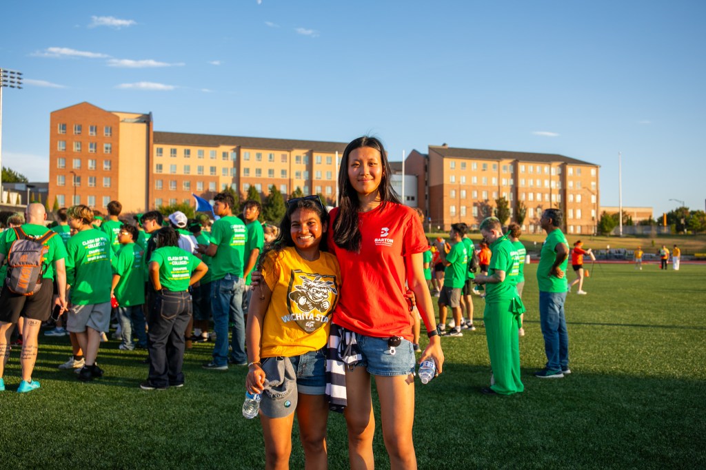 Two participants posing together on a grassy field during a campus competition, with residence halls and other attendees in the background.