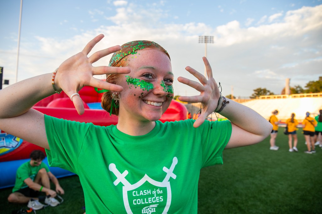 Participant wearing a green “Clash of the Colleges” T-shirt, with green glitter on their face and hands, posing on a field during an outdoor campus competition.