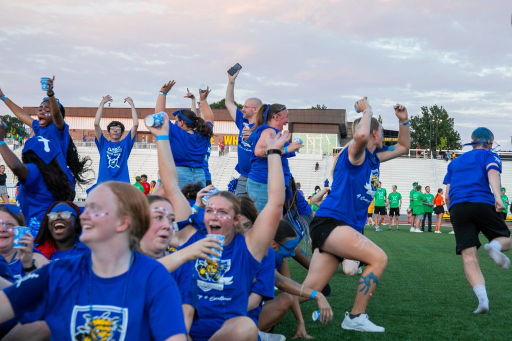 Participants in blue team shirts cheering and celebrating together on a field during a Clash of the Colleges event.