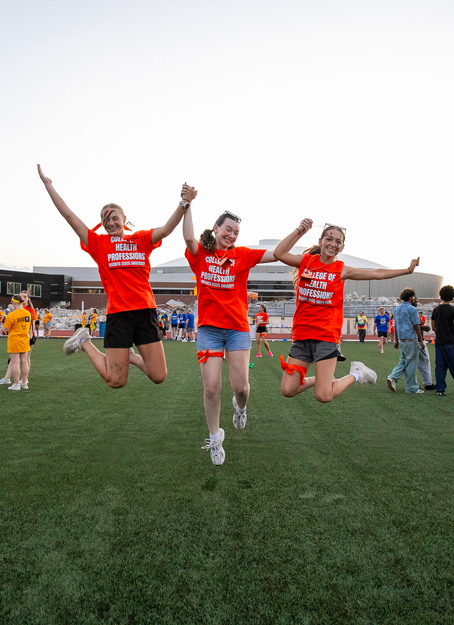 Three participants in matching red team shirts jumping in the air on a field during a campus event.