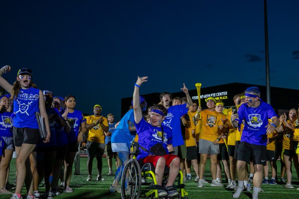 Participant in a wheelchair at the front of a group of cheering students in blue and gold team shirts on a lit outdoor field at night.
