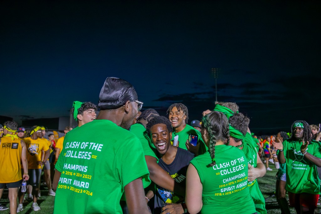 Group of participants in green team shirts gathering together on a field during an evening Clash of the Colleges event.