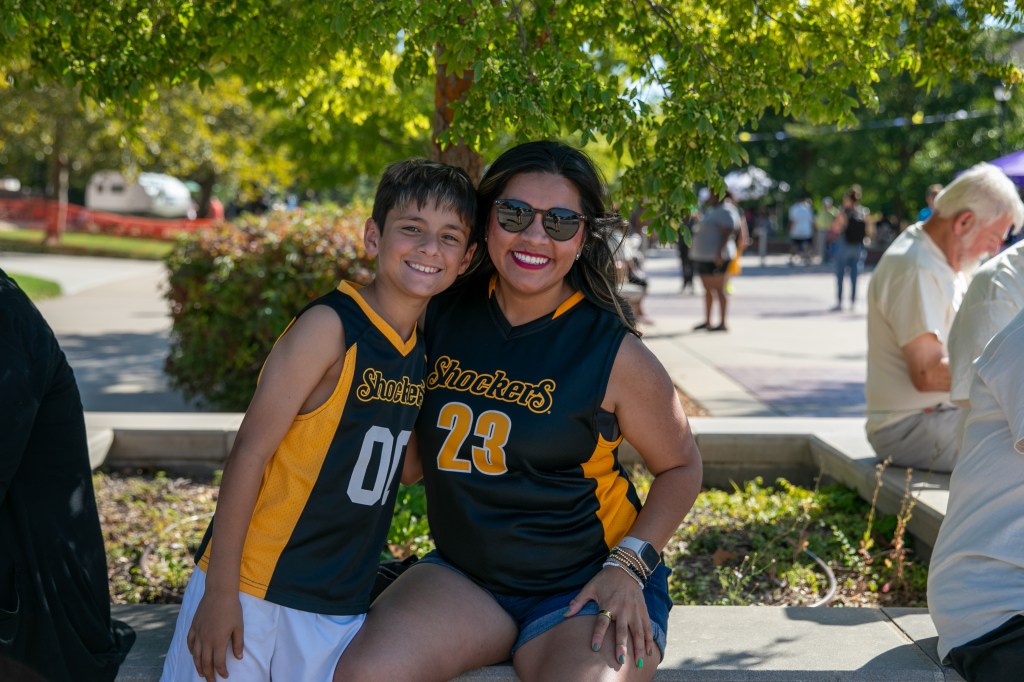 Two people wearing black-and-gold Wichita State Shockers jerseys posing together outdoors beneath leafy trees.