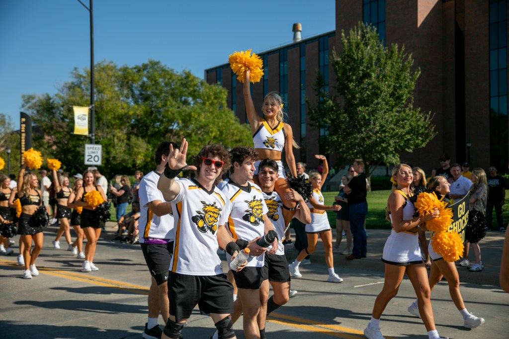 Wichita State students and cheerleaders walking in a parade, with one cheerleader raised on a participant’s shoulders holding a gold pom-pom.
