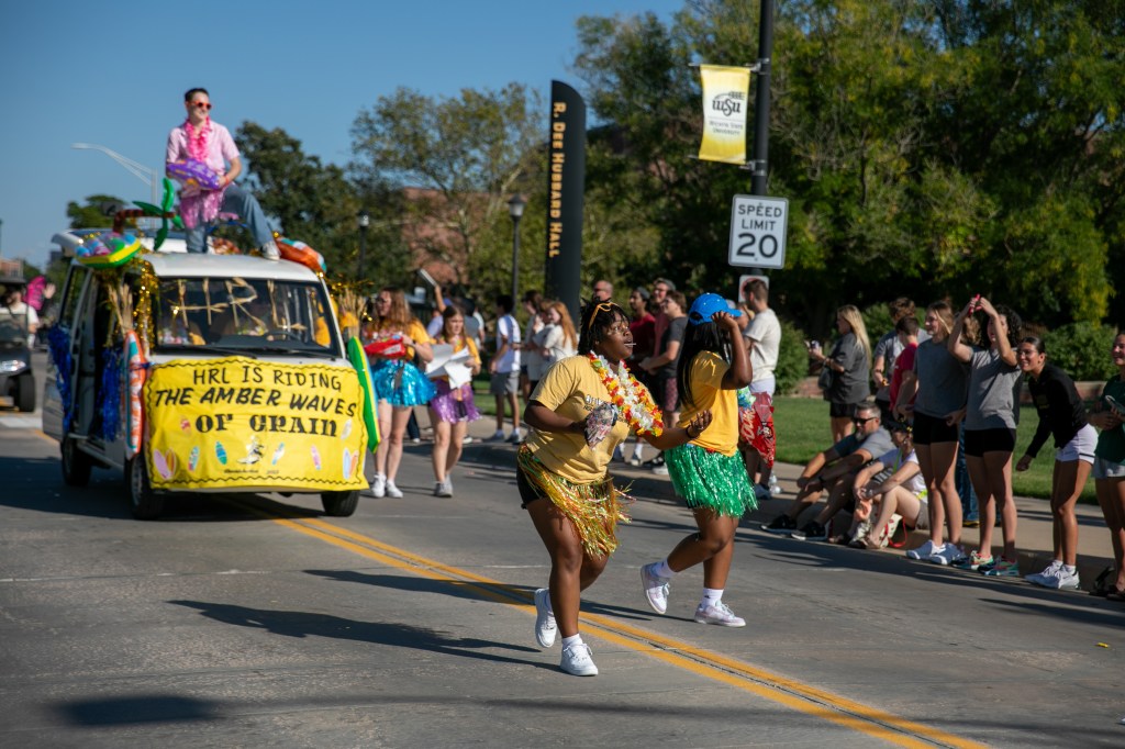 Parade participants dancing in the street alongside a decorated vehicle, with spectators gathered along the roadside.