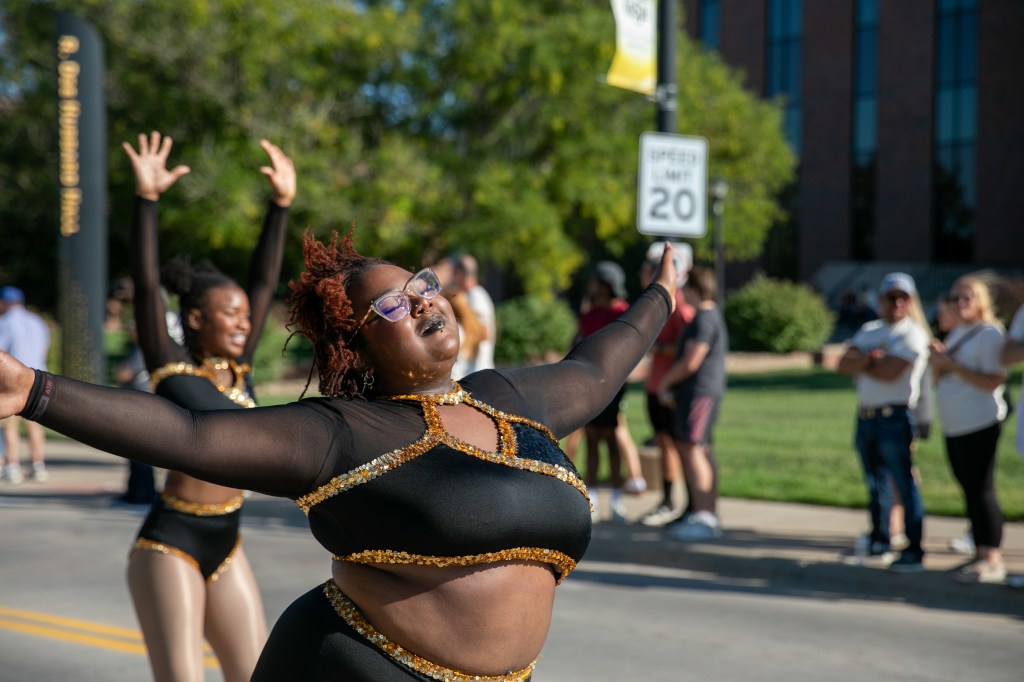 Performer in a black-and-gold costume posing with outstretched arms during an outdoor campus celebration, with spectators in the background.