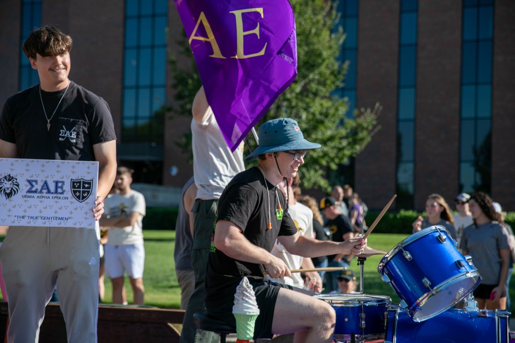 Drummer performing at an outdoor campus event, seated behind blue drums while holding drumsticks, with a purple flag visible nearby.