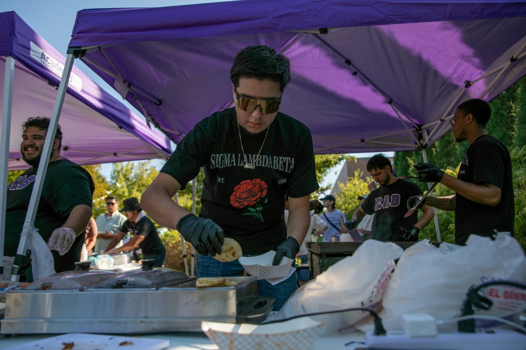 Person preparing food at an outdoor event booth beneath a purple canopy tent.