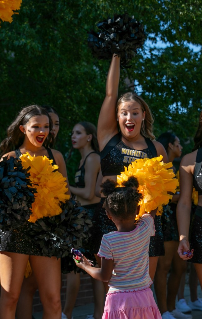 Wichita State cheerleaders holding black and gold pom-poms while interacting with a child during an outdoor campus event.