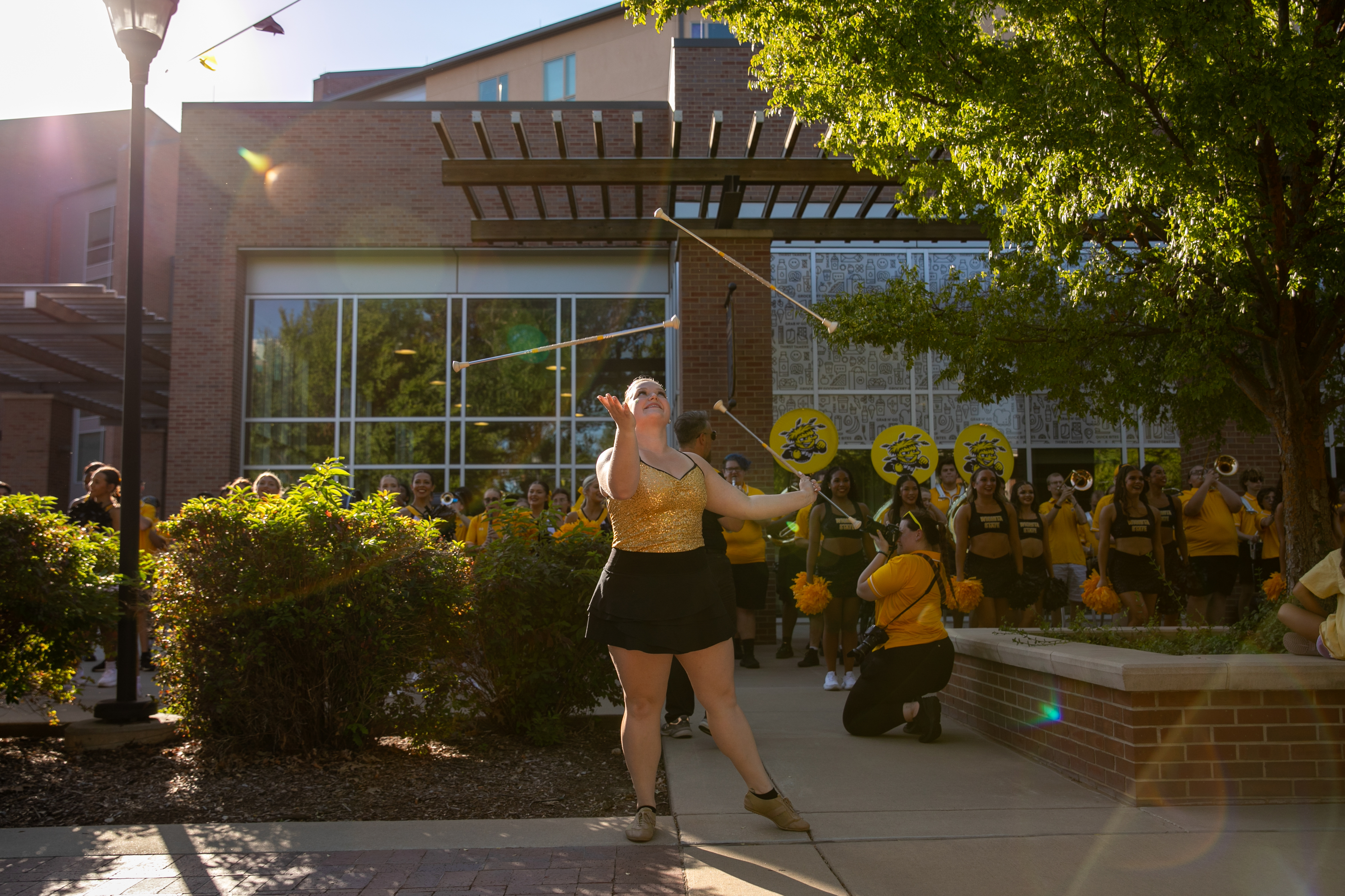 Person performing a baton-twirling routine on a campus walkway while a crowd gathers nearby and Wichita State spirit signs are visible in the background.