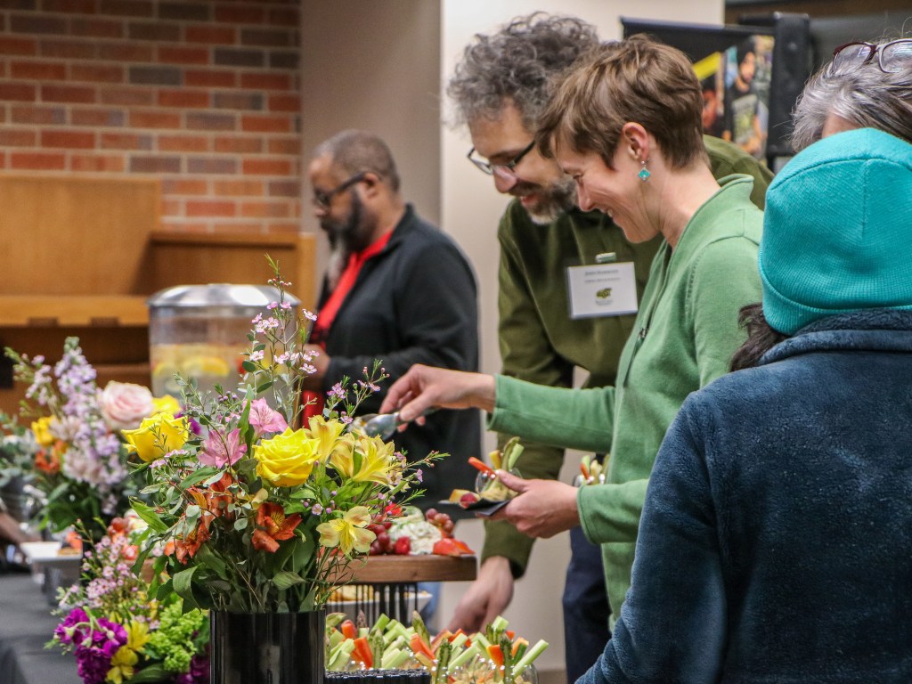 Participants selecting food from a buffet-style table decorated with colorful flower arrangements during an indoor reception.