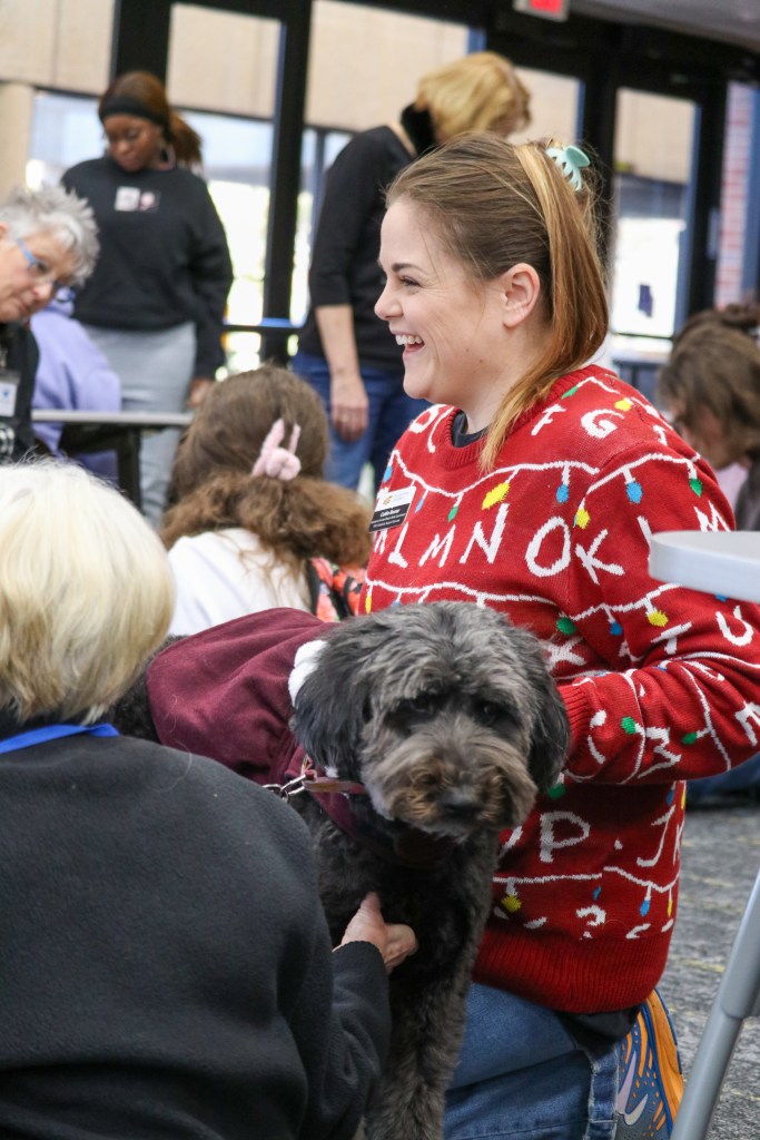 Attendees gathered indoors around a black dog as part of a therapy or pet engagement activity.