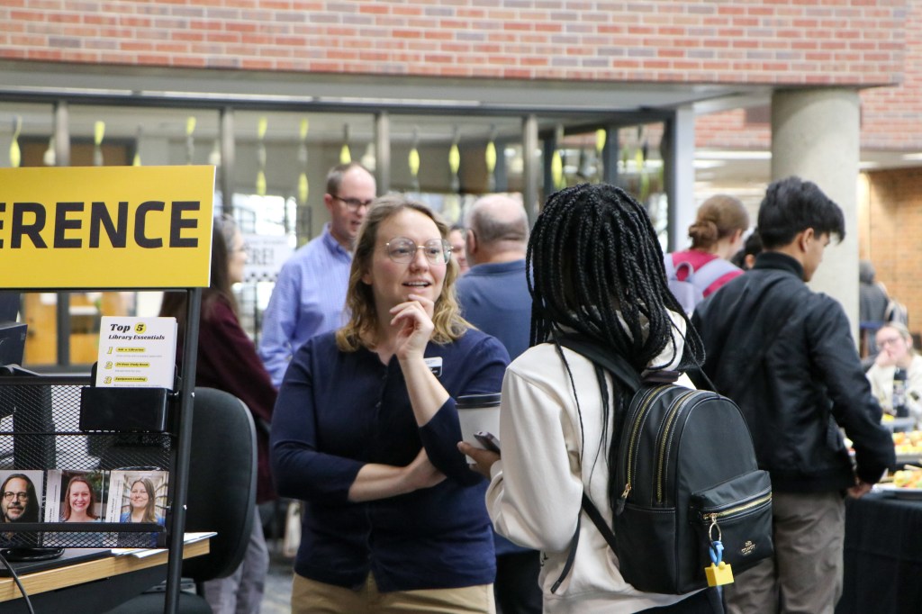 Attendees conversing at an indoor information fair, with a display sign and resource table in the foreground.