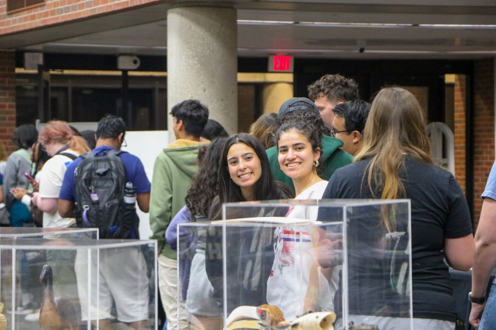 Attendees gathered indoors around display cases at a campus event, with two participants looking toward the camera in the foreground.